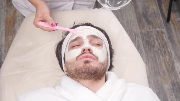 Έννοια του spa για τους άνδρες. Ένας νεαρός άνδρας εφαρμόζεται ένα καλλυντικό μάσκα στο πρόσωπο σε ένα σαλόνι ομορφιάς — Αρχείο Βίντεο