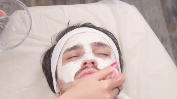 Conceito de spa para homens. Um jovem é aplicado uma máscara cosmética no rosto em um salão de beleza — Vídeo de Stock