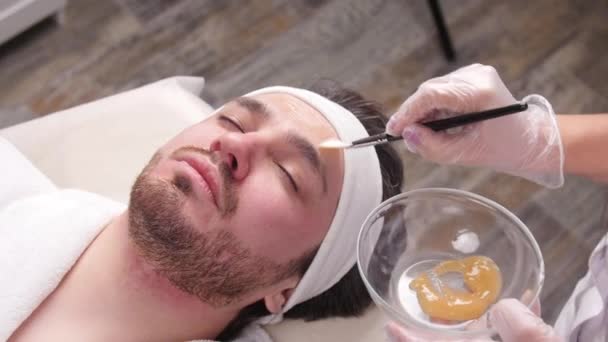 Концепция спа для мужчин. Молодой человек надевает косметическую маску на лицо в салоне красоты — стоковое видео