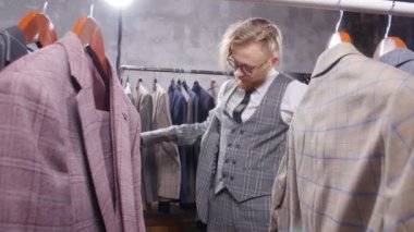 Alışveriş ve moda konsepti - Alışveriş merkezinde veya giyim mağazasında ceketi seçen ve deneyen genç sakallı adam