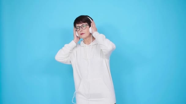 Mulher de meia-idade em óculos e uma camisola branca escuta sua música favorita com fones de ouvido em um fundo branco. Conceito de assinatura de rádio e música online — Vídeo de Stock