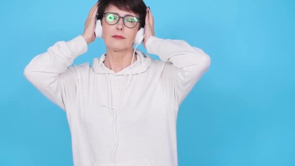 Donna di mezza età in occhiali e un maglione bianco ascolta la sua musica preferita con le cuffie su sfondo bianco. Concetto di abbonamento radio e musica online — Video Stock