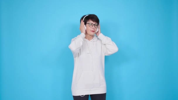 Liefde voor muziek concept. Rijpe vrouw streamt muziek in een koptelefoon op een blauwe achtergrond — Stockvideo
