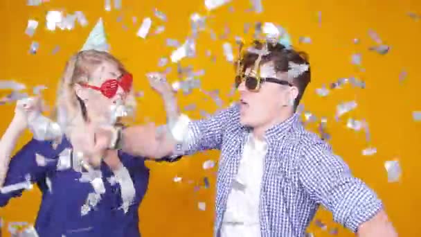 Concepto de vacaciones y cumpleaños. Joven pareja feliz bailando en sombreros sobre fondo naranja con confeti — Vídeos de Stock