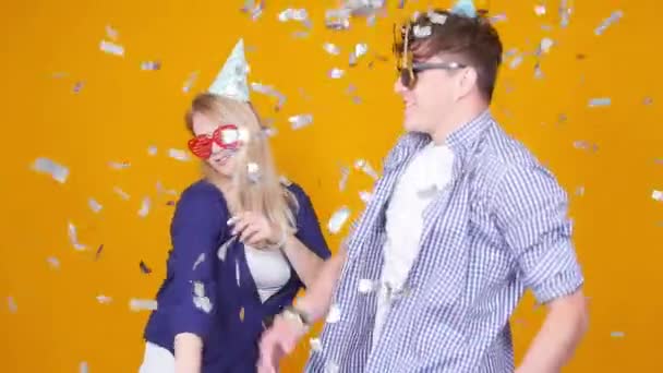 Concept de vacances et d'anniversaire. Jeune couple heureux dansant dans des chapeaux sur fond orange avec des confettis — Video