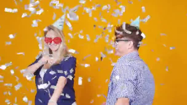 Concepto de vacaciones y cumpleaños. Joven pareja feliz bailando en sombreros sobre fondo naranja con confeti — Vídeos de Stock