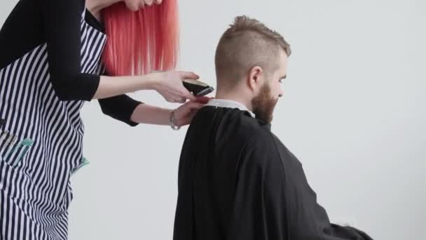 Peluquería femenina pelirroja joven está cortando el pelo del hombre barbudo cliente — Vídeo de stock