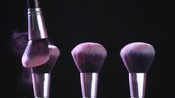 Conceito de beleza. Escovas cosméticas com pó cosmético rosa espalhando no fundo preto em câmera lenta — Vídeo de Stock