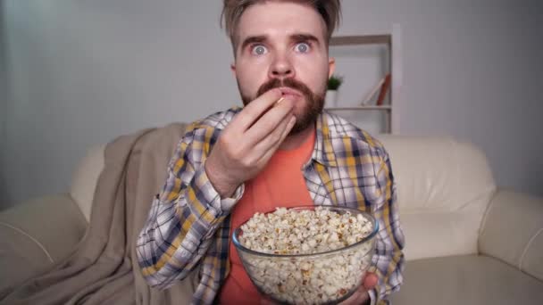 Концепція домашнього кіно. Шокований і наляканий чоловік дивиться телевізор і їсть попкорн — стокове відео