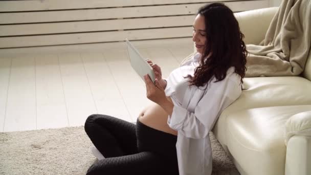 Молодая беременная женщина дома смотрит на планшетный компьютер — стоковое видео