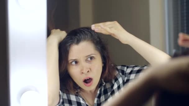 Νεαρή απογοητευμένη γυναίκα κοιτιέται στον καθρέφτη και βγάζει μια γκρίζα τρίχα από το κεφάλι της. Concept φωτογραφία της νεαρής γυναίκας να γερνάει — Αρχείο Βίντεο