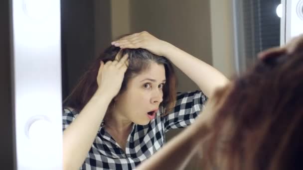 Разочарованная молодая женщина смотрит в зеркало и выщипывает седые волосы из головы. Концепция фото молодой женщины стареет — стоковое видео