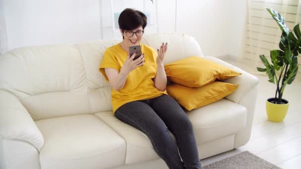 Koncept sebeizolace. Žena středního věku sedí na pohovce v obývacím pokoji a mluví prostřednictvím videohovoru. — Stock video