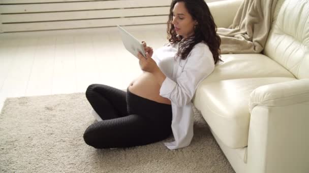 Молода вагітна жінка вдома дивиться на планшетний комп'ютер — стокове відео