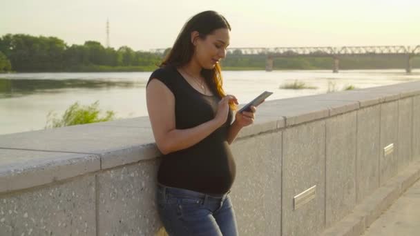 Νεαρή έγκυος γυναίκα που χρησιμοποιεί ένα έξυπνο τηλέφωνο στέκεται σε ένα πάρκο — Αρχείο Βίντεο