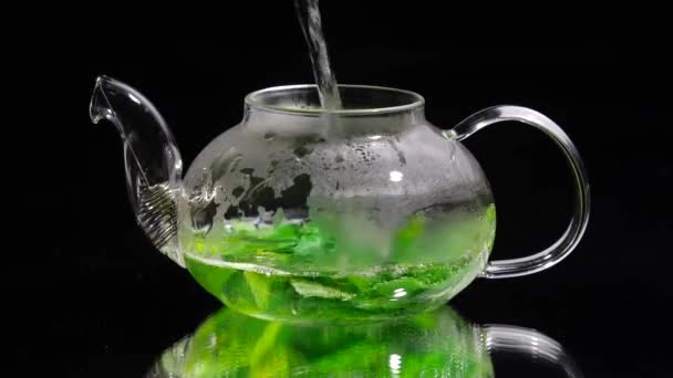 Verser de l'eau chaude dans une théière en verre sur un fond noir, thé à la menthe verte — Video