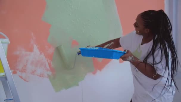 Koncepcja renowacji mieszkania. Uśmiechnięta Afroamerykanka przemeblowuje swój dom trzymając wałek malarski pokryty pomarańczową i zieloną farbą — Wideo stockowe