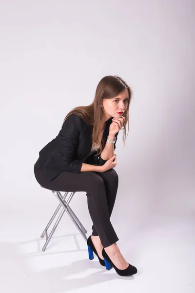 Po celé délce studiový portrét mladé ženy v černém obleku, bot na podpatku, sedí v křesle na bílém pozadí — Stock fotografie