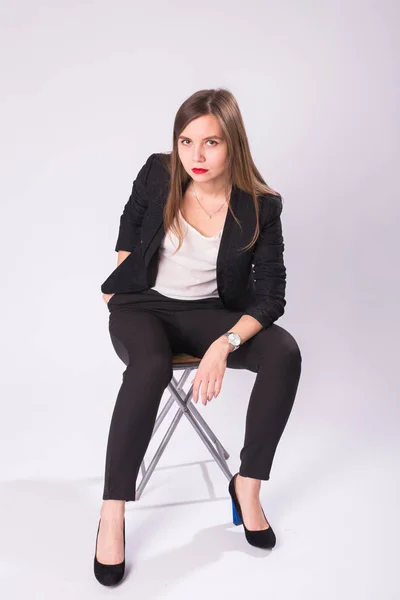 Retrato de estudio de larga duración de una joven mujer de negocios en traje negro, zapatos de tacón alto, sentada en silla sobre fondo blanco — Foto de Stock
