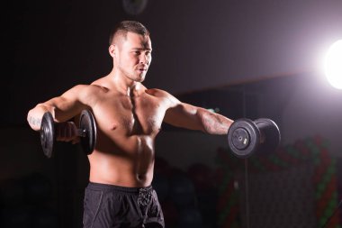 Güçlü vücut geliştirmeci dumbbells ile egzersizleri yapıyor. Koyu arka plan üzerinde çıplak gövde ile güçlü erkek. Güç ve motivasyon.