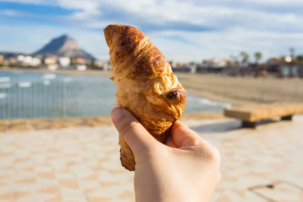 Croissant na mão contra o pano de fundo de uma bela praia — Fotografia de Stock