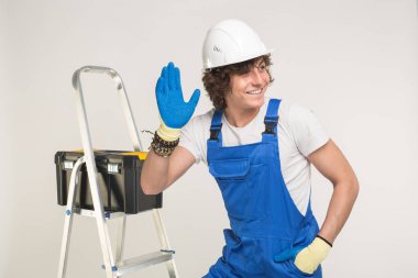 Stüdyo portre yakışıklı Builder beyaz kask ve genel olarak elini sallayarak ve gülen mavi.