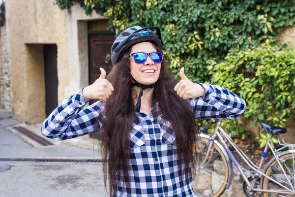 Γυναίκα στο κράνος και γυαλιά ηλίου δείχνουν αντίχειρες στο παρασκήνιο ποδηλάτων και Οδός — Φωτογραφία Αρχείου