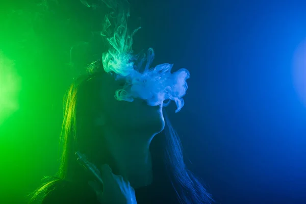 Portrait of young woman in neon blue and green smoke with vape or e-cigarettes — Stock Photo, Image