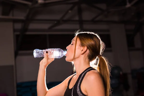 フィットネスの若い女性は、ボトルから水を飲む。ジムのトレーニングから休憩を取る筋肉の若い女性. — ストック写真