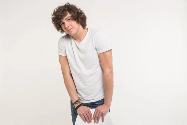Stilig ung man med lockigt hår stående i vit t-shirt bakom en stol på vit bakgrund — Stockfoto
