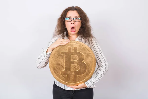 Mujer de negocios sorprendida con la boca abierta mirando cámara y sosteniendo gran bitcoin de oro sobre fondo blanco — Foto de Stock