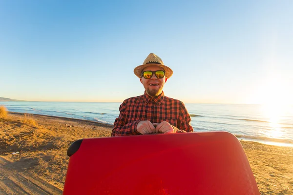 Viajar, vacaciones y concepto de personas - hombre feliz en gafas de sol y sombrero llevando su maleta roja cerca del mar — Foto de Stock