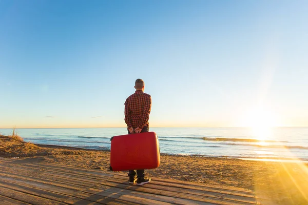 Concepto de naturaleza, viajes y personas: vista trasera del hombre sentado en una maleta roja cerca del mar y mirando al atardecer — Foto de Stock