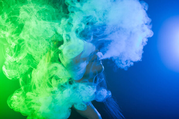 Portrait of young woman in neon blue and green smoke with vape or e-cigarettes