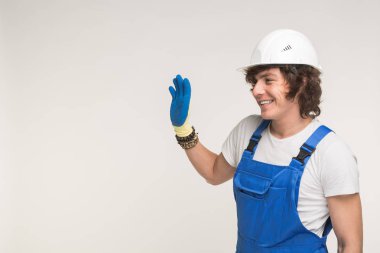 Stüdyo portre yakışıklı Builder beyaz kask ve genel olarak elini sallayarak ve gülen mavi