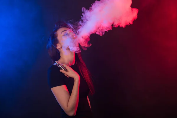 Young woman in neon light and smoke of e-cigarettes or vape on dark background — Stock Photo, Image