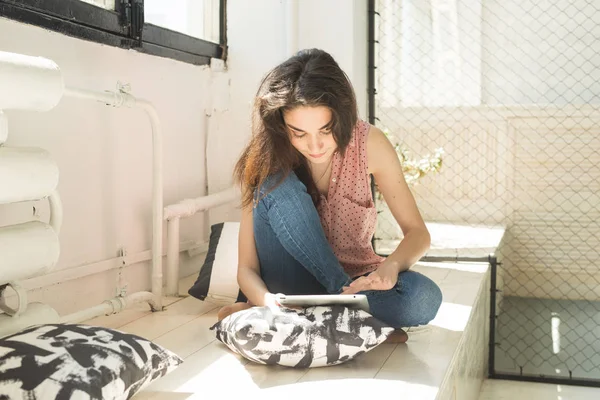 Tecnologia e concetto di persone - giovane donna seduta sul davanzale di legno con tablet e cuscini — Foto Stock