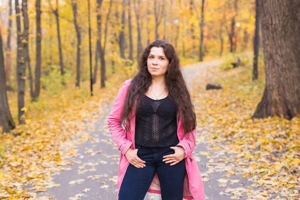 Season, fashion and people concept - plus size woman in blue pants and pink jacket standing in autumn forest — Stock Photo, Image