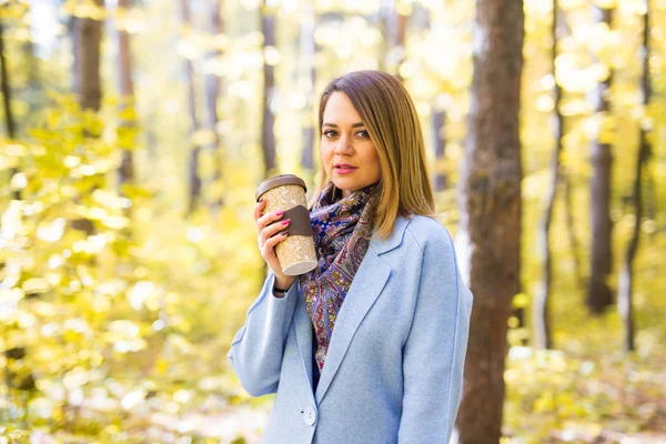秋には、自然、人々 の概念 - 公園一杯のコーヒーで、青いコートに立っての若いブルネットの女性 — ストック写真