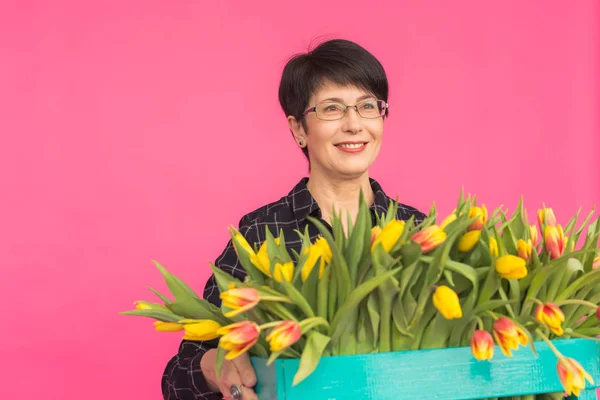 Mooie middelbare leeftijd vrouw met gele tulpen op roze achtergrond. Floristiek, vakantie en geschenken concept — Stockfoto