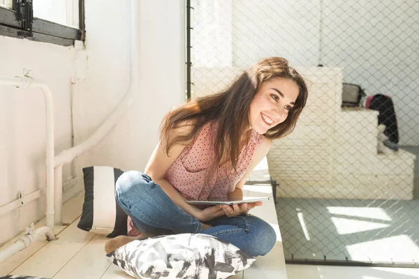 Koncepcja ludzie, technologia i wnętrze - szczęśliwa młoda kobieta siedzi na podłodze z komputera typu tablet z poduszki wokół — Zdjęcie stockowe