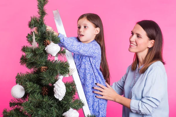 Christmas and holiday concept - Portrait of smiling woman with her little daughter decorating fir tree over pink background — Stock Photo, Image