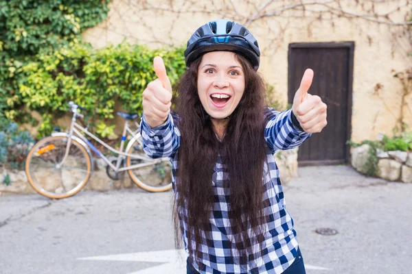 Νεαρή γυναίκα χαμογελαστή με κράνος και γυαλιά ηλίου δείχνουν αντίχειρες στο παρασκήνιο σοκάκι και ποδηλάτων — Φωτογραφία Αρχείου