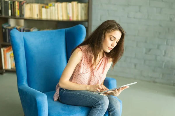 Technológiák, emberek koncepció - fiatal nő ül egy széken, és nézi, a tablet vagy szörfözés a háló, és mosolyogva — Stock Fotó