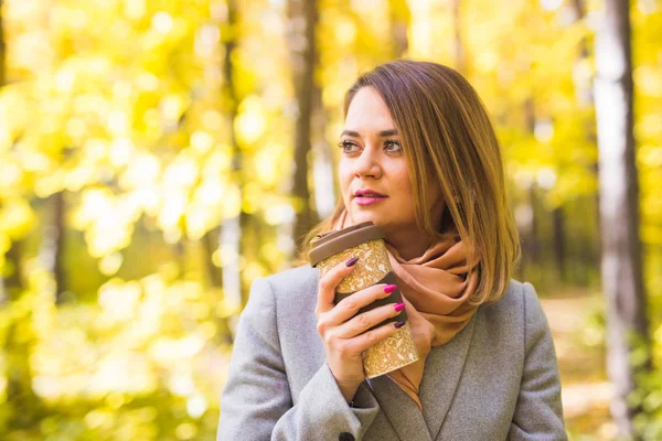 Herfst, natuur, mensen concept - jonge vrouw in een blauwe jas staan in het park op een achtergrond van bomen met een kopje koffie. — Stockfoto