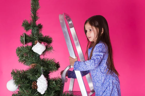 Weihnachts- und Urlaubskonzept - ein kleines Mädchen schmückt Weihnachtsbaum auf rosa Hintergrund — Stockfoto