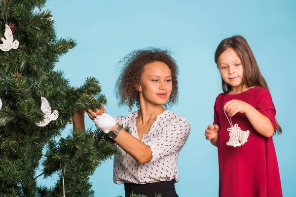 Helgdagar, familj och jul koncept - söt liten flicka med blandad ras mamma dekorerar en julgran på blå bakgrund — Stockfoto