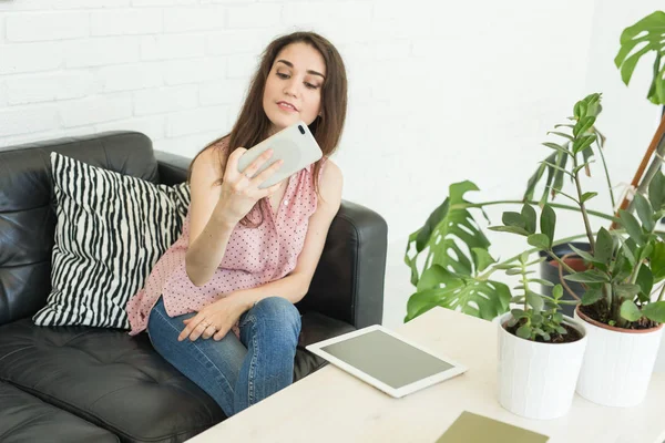 Concept de technologie, d'internet et de personnes - jeune blogueuse brune faisant des photos à l'aide d'un smartphone et allongée sur un canapé à la maison — Photo