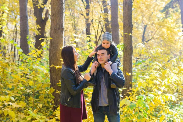 Family, autumn, people concept - young family walking in autumn park. Daughter sitting on dads shoulders