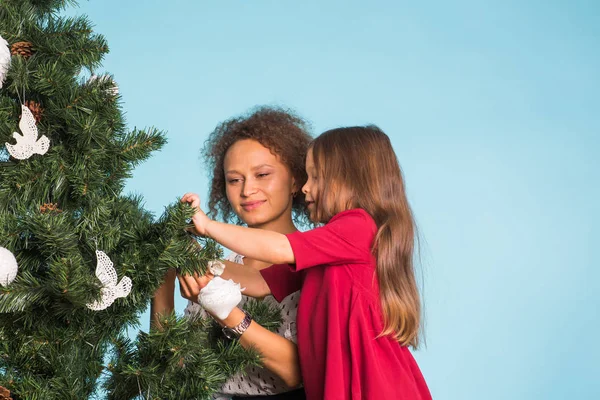 Holidays, family and christmas concept - pretty little girl with mixed race mother are decorating a christmas tree on blue background
